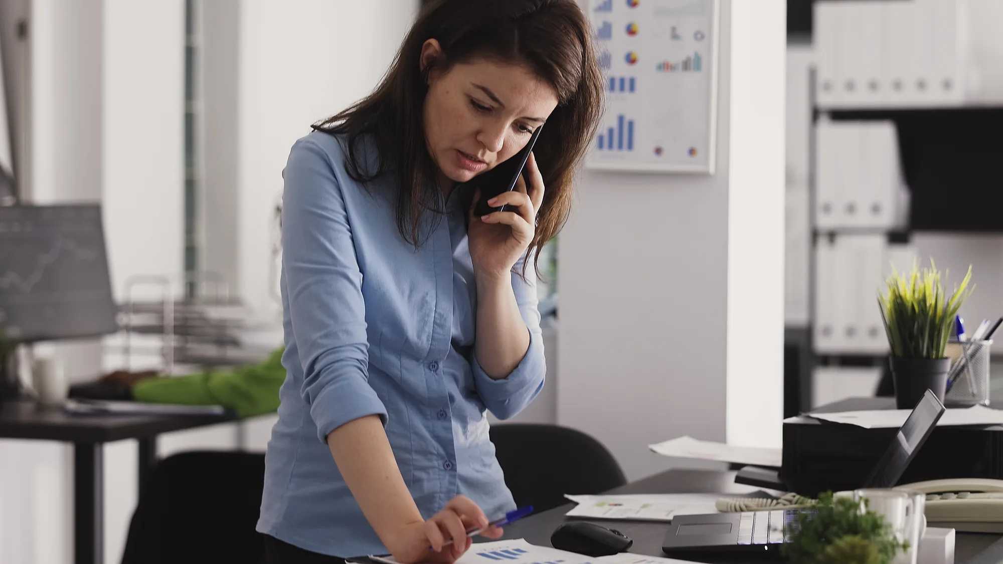 A woman on the phone leaning over paperwork at a desk, pen in hand