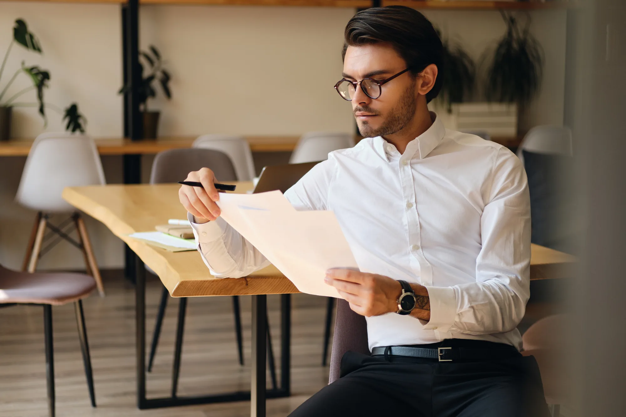 A man in glasses reading two printed sheets at a wooden table in a bright office
