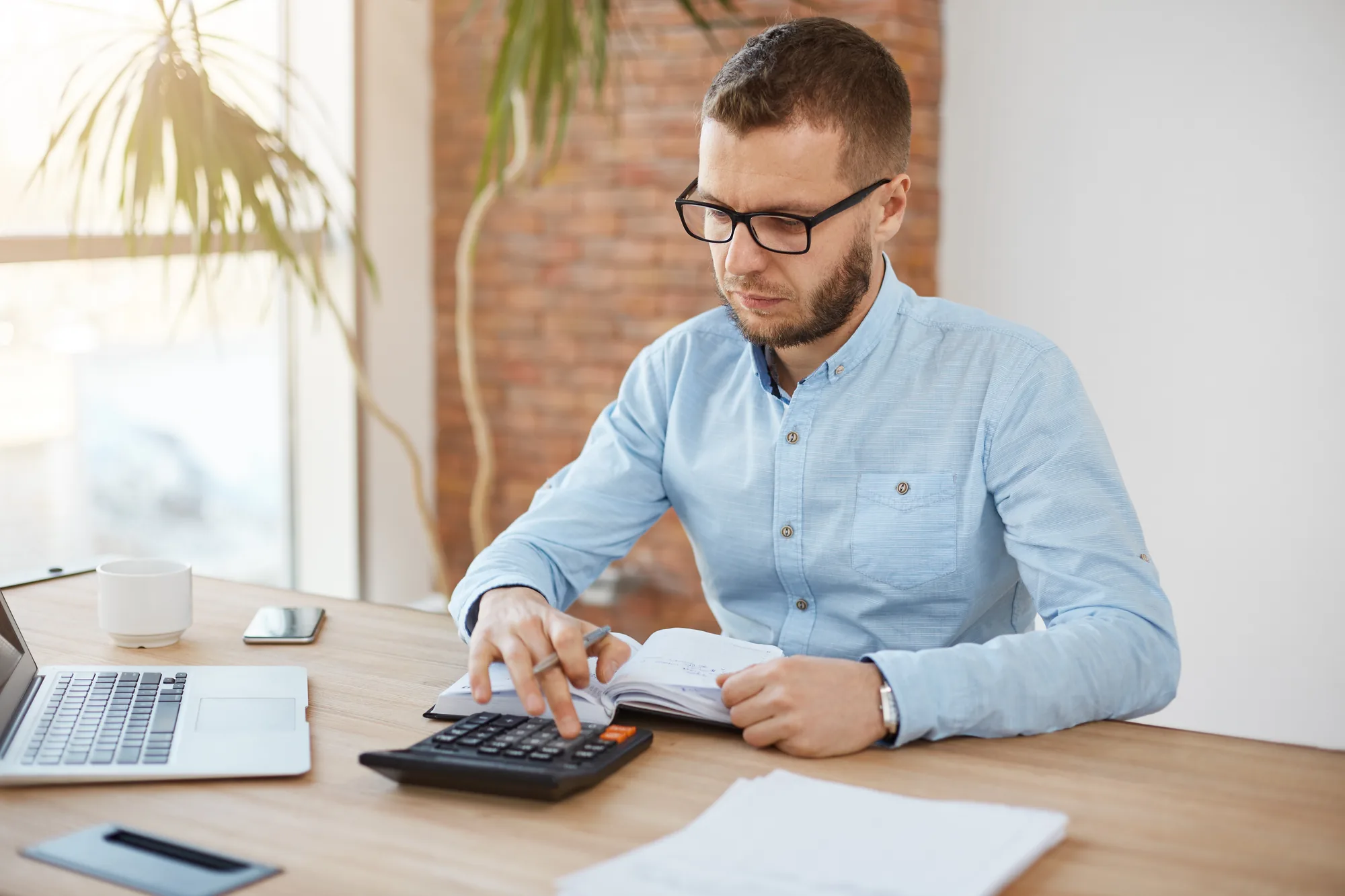 Man in glasses keying a calculator beside an open laptop and notebook in a bright brick-walled office