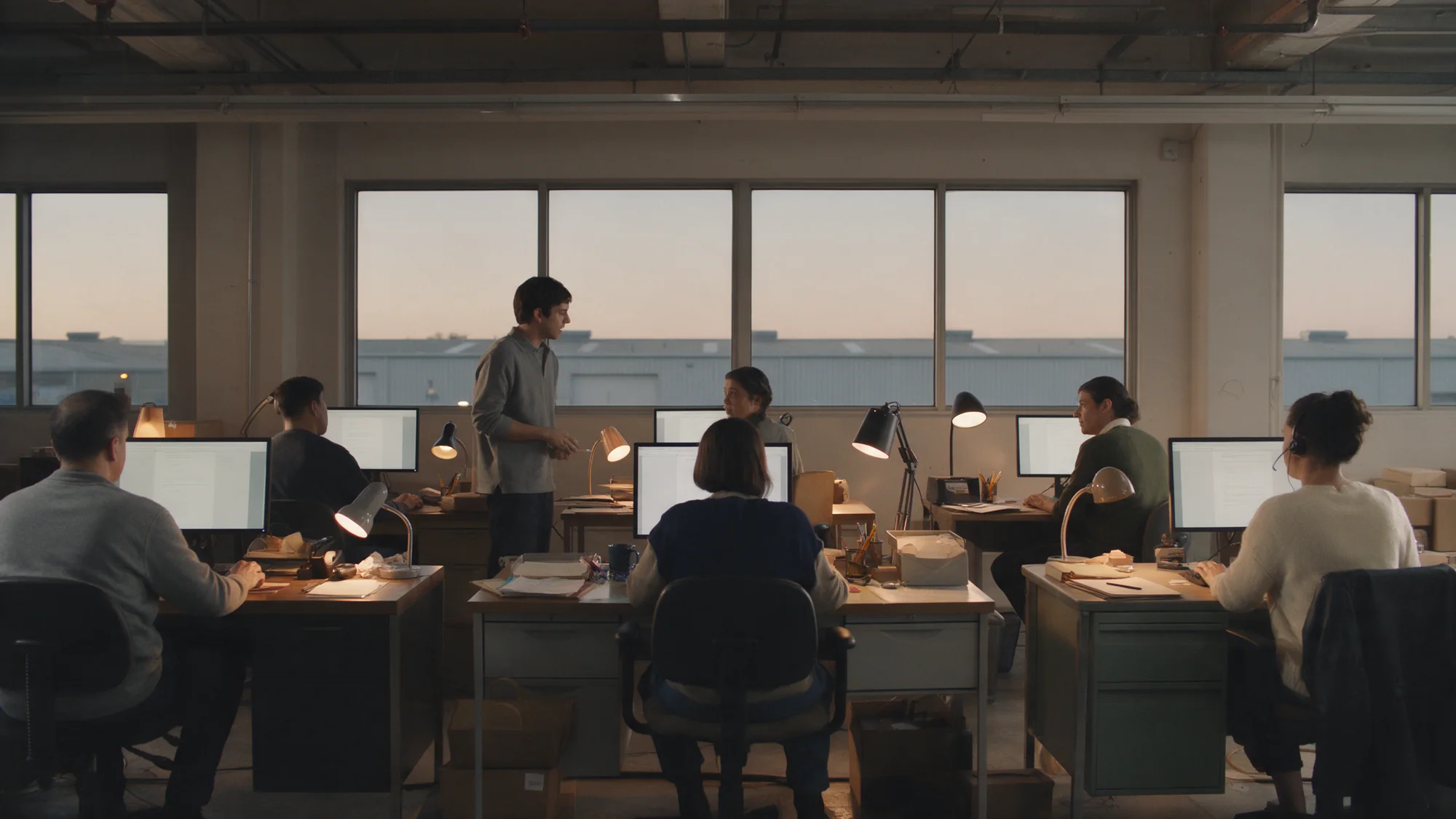 An operations floor at dusk with six desks, people at monitors and warm desk lamps
