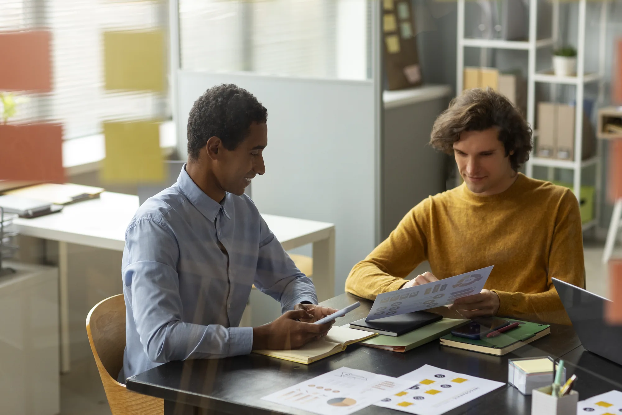 Two colleagues at a desk going through printed pages beside a laptop and a tablet