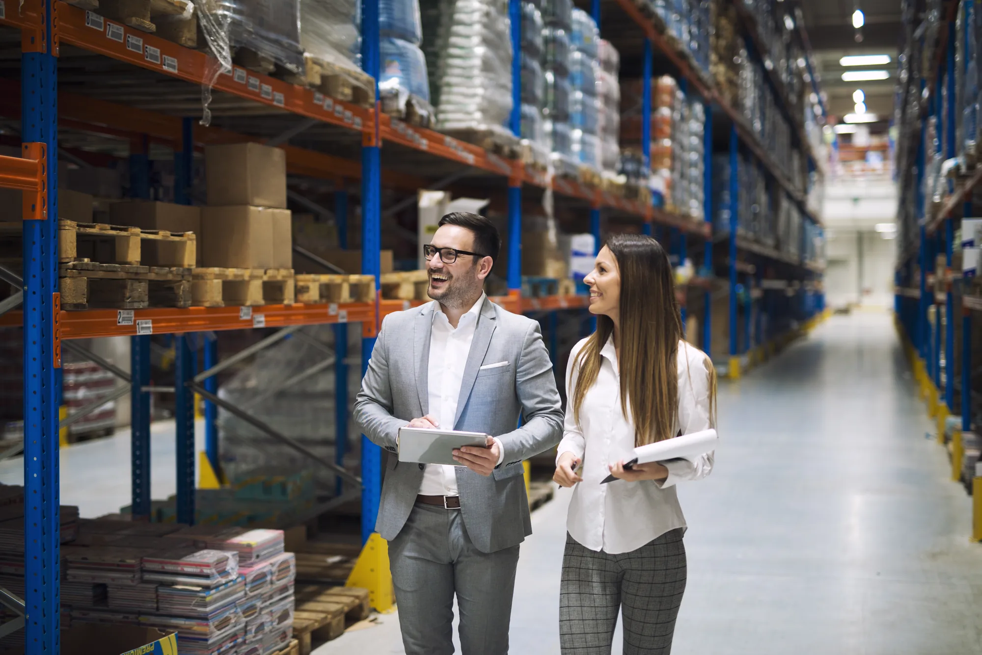 A man in a grey suit carrying a tablet and a woman carrying a folder walking and talking down a long warehouse aisle between tall racking